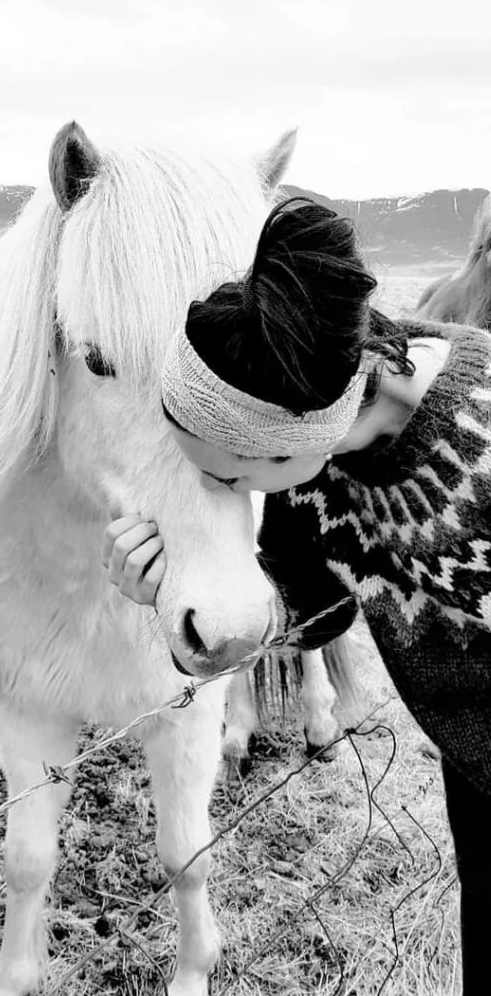 Das Bild scheint in Schwarzweiß aufgenommen zu sein, wobei Texturen und Kontraste vor einem gedämpften Hintergrund betont werden, der an eine Freiluftumgebung mit Anklängen an eine ländliche oder ländliche Umgebung erinnert. Der Fokus liegt auf einer Person und einem Pferd, die einen Moment stiller Zuneigung teilen.